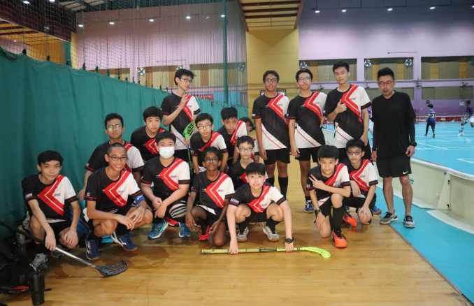 Youth floorball team in black and red uniforms posing with sticks inside an indoor sports hall.